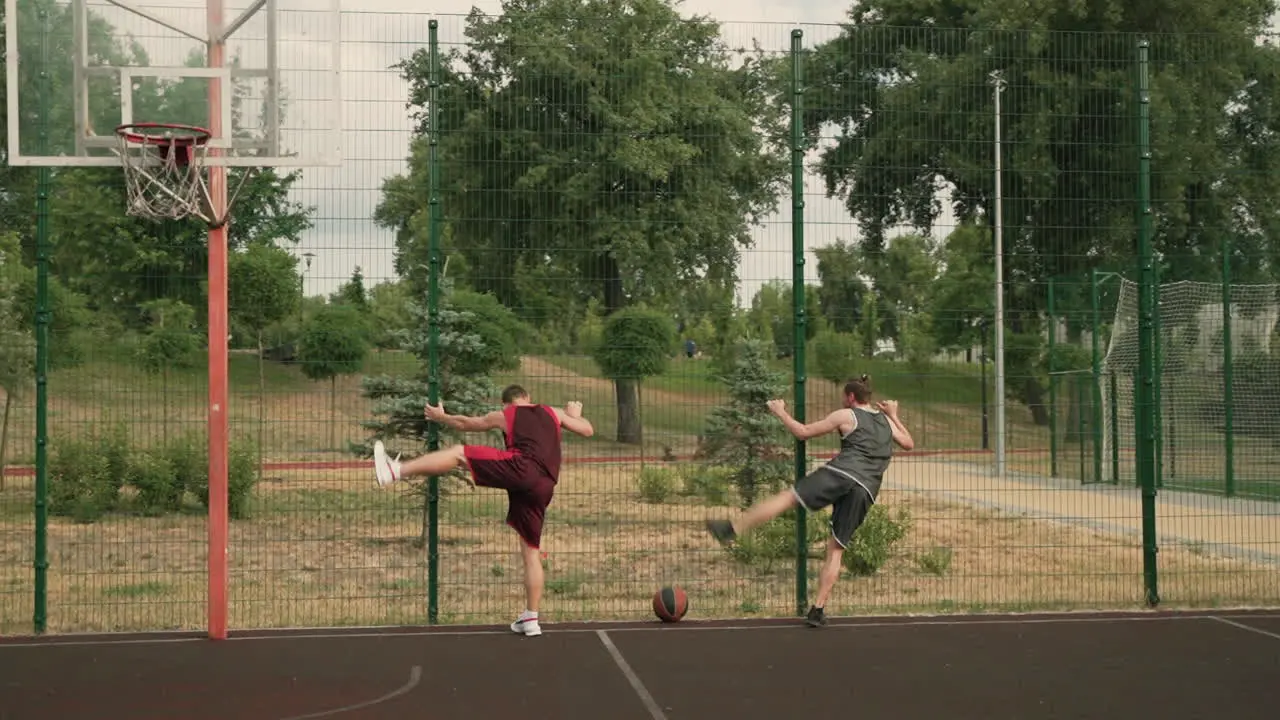 Two Male Basketball Players Stretching Their Legs Standing And Leaning Against A Metal Fence In An Outdoor Basketball Court