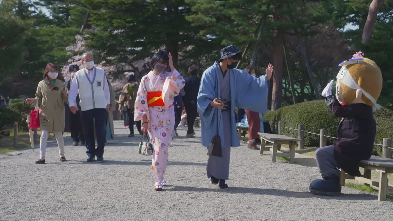 Young asian Japanese masked couple in kimono walking in Kenrokuen Garden and waving to a local mascot litakun