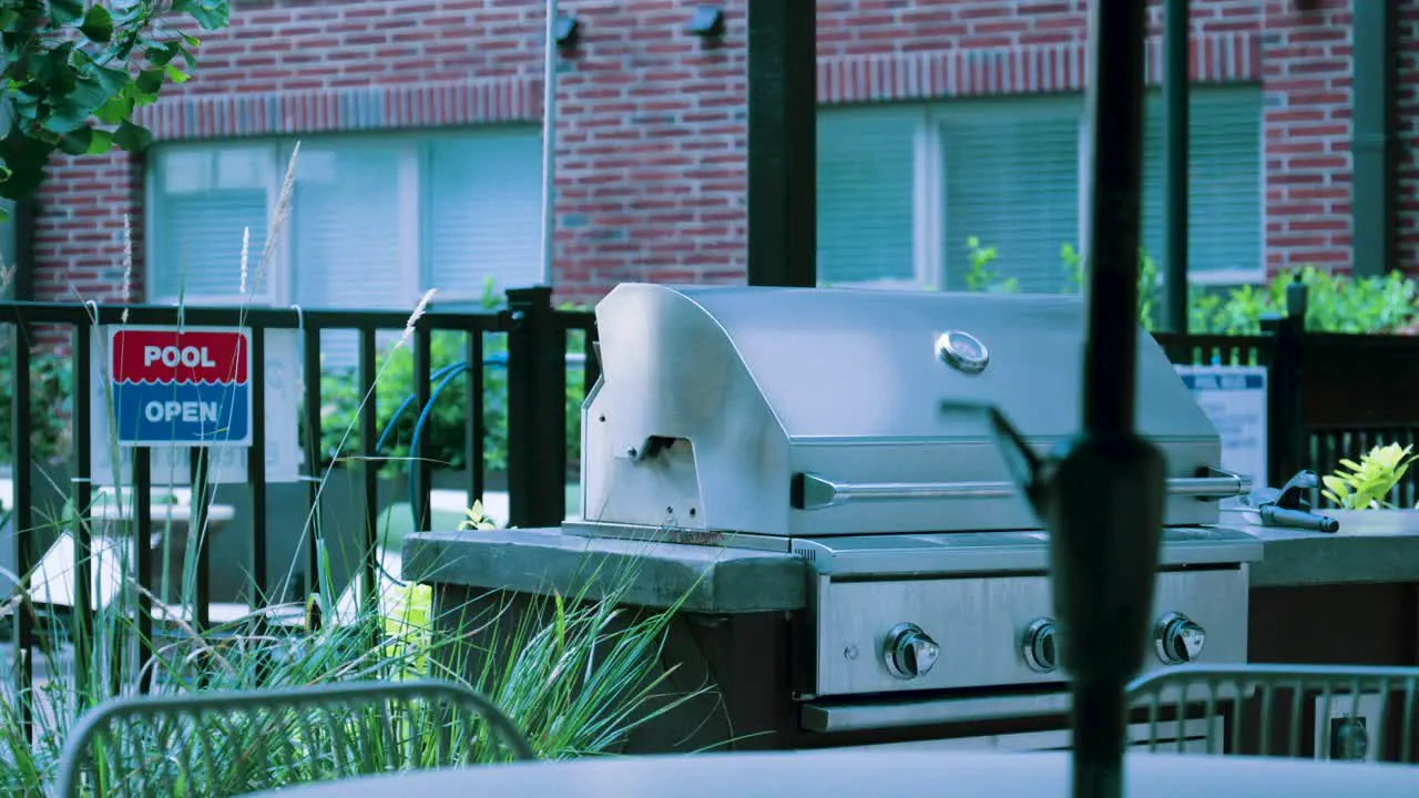 Heat waves rise from a stainless steel BBQ grill near an open pool sign and wrought iron gate backed by a brick building and with a picnic table in the foreground