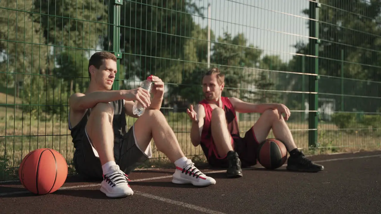 Two Male Basketball Players Chatting While Sitting And Leaning Against A Metal Fence In An Outdoor Basketball Court