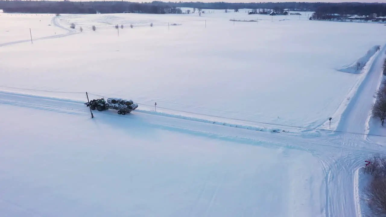 Aerial view tractor trailer carrying rolled bales across snowy winter rural agricultural countryside