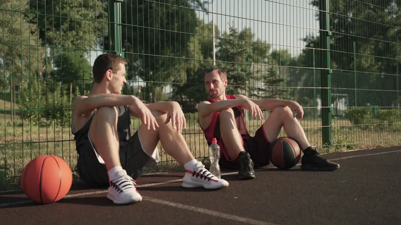 Two Happy Basketball Players Chatting While Sitting And Leaning Against A Metal Fence In An Outdoor Basketball Court