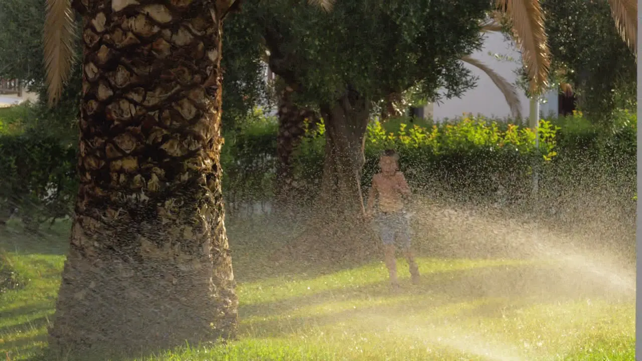 Child running on the lawn and getting wet from water sprinkler