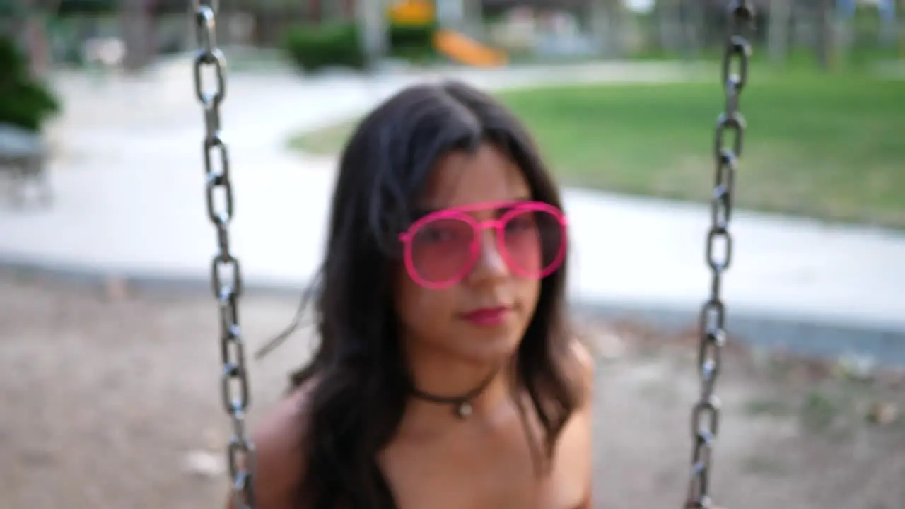 A beautiful hipster fashion model wearing vintage clothing and retro pink sunglasses swinging on a playground and having fun SLOW MOTION