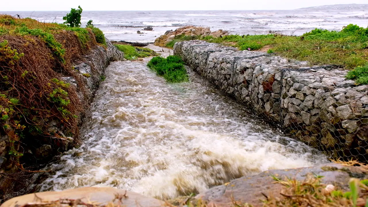 Rainwater runoff gushing out of stormwater drain with gabion walls toward ocean