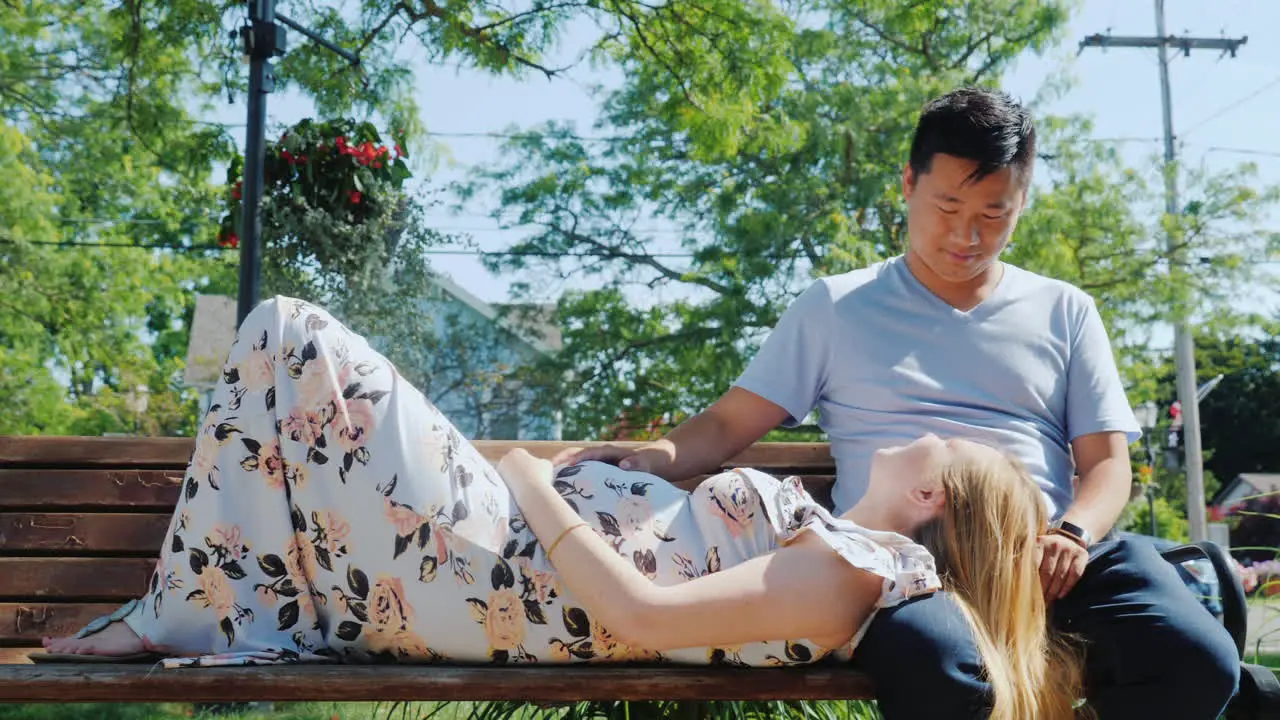 A Pregnant Woman With Her Husband Are Resting In A Park On A Bench In A Typical Suburb Of Usa