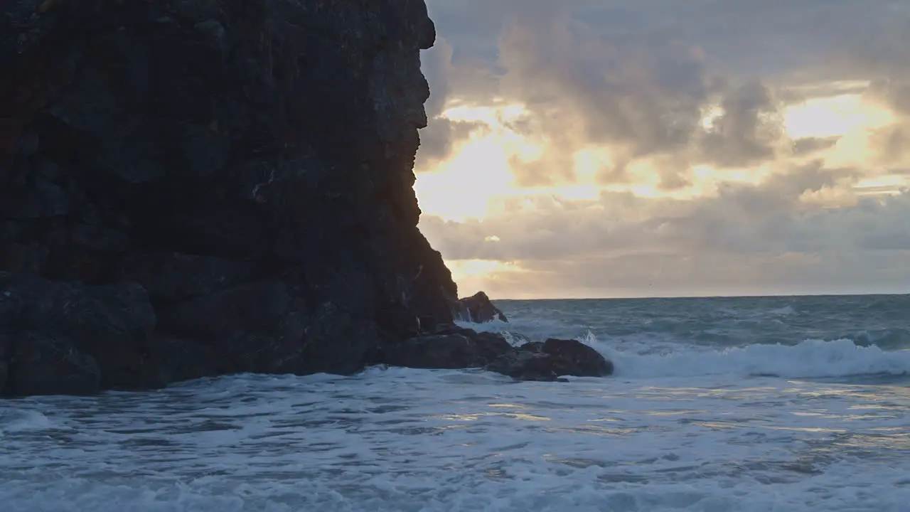 Waves crash on cliffs silhouetted by blazing sun through clouds