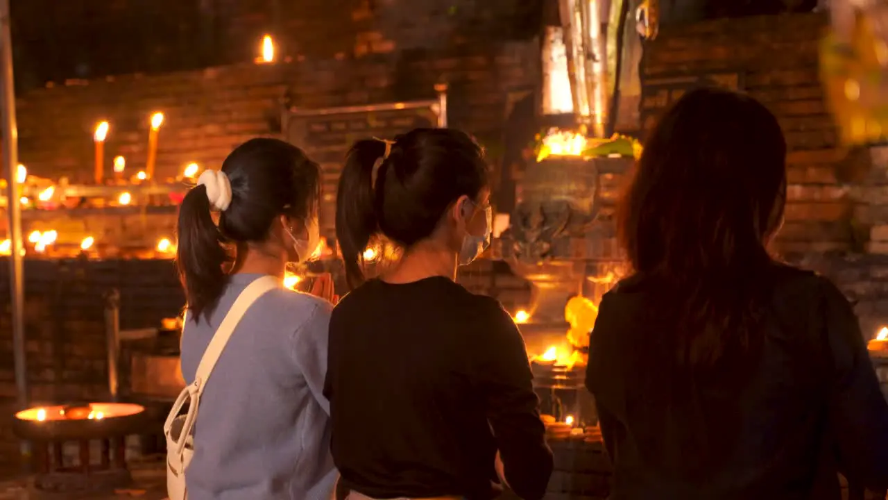 Back of three young female praying at Buddhist temple lit up with candles