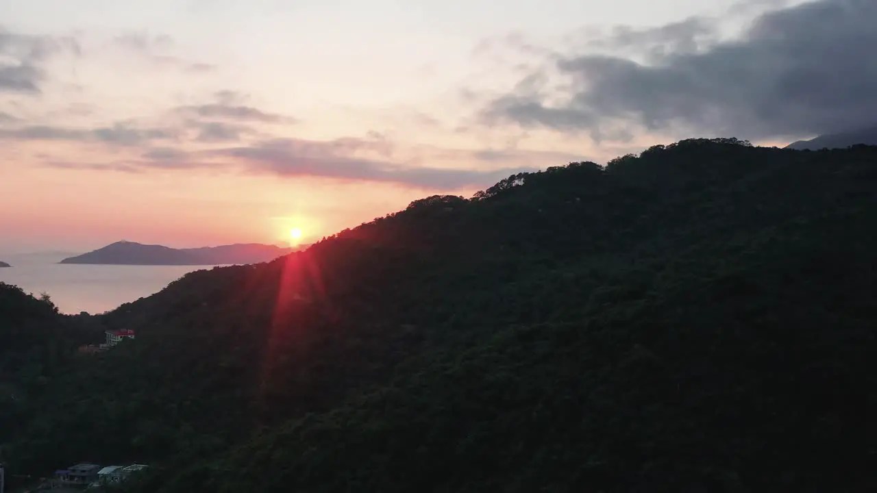 Aerial truck left of sunset sunbeams behind rainforest hills and village in Pui O bay sea in background Lantau Island Hong Kong China