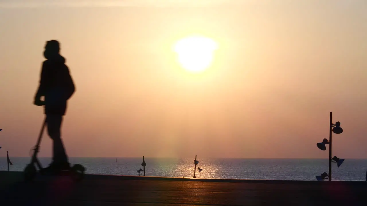 Man on electric scooter briefly stops to admire pink ocean sunrise exits left