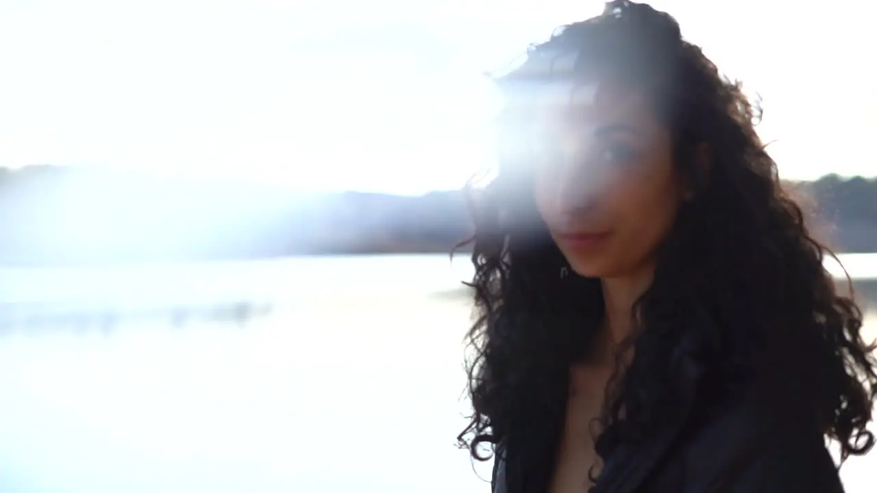 Pretty young woman with thick long curly brunette hair at a lake by the forest and the bright sunshine behind her orbit shot with light leak lens flare