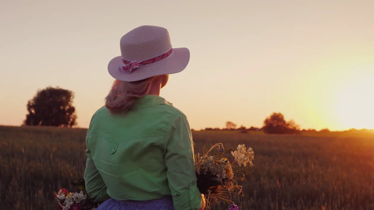 A Romantic Woman With A Hat And A Bouquet Of Wildflowers Admires The Sunset Over The Wheat Field