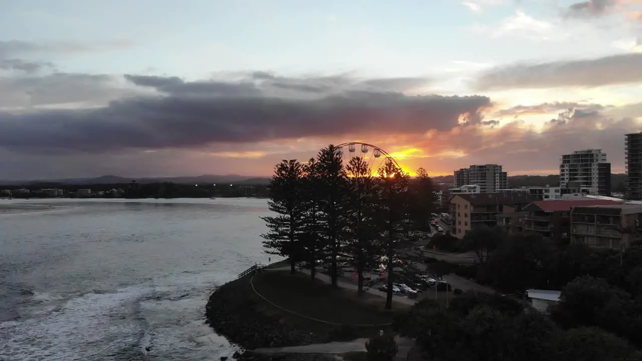 A small ferris wheel sits hidden by treats next to a beautiful beach behind lies a small beach city where a sun burning through the darkened clouds descends behind tall mountains in the distance