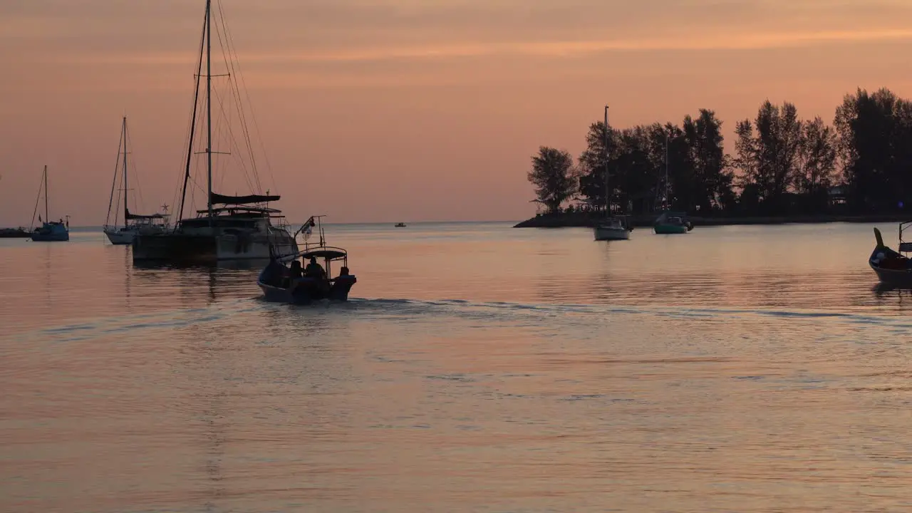 Tropical sunset scenery capturing shimmering sky reflection on calm water surface with fishing boat cruising forward and luxurious yacht anchored in the bay Langkawi island Malaysia