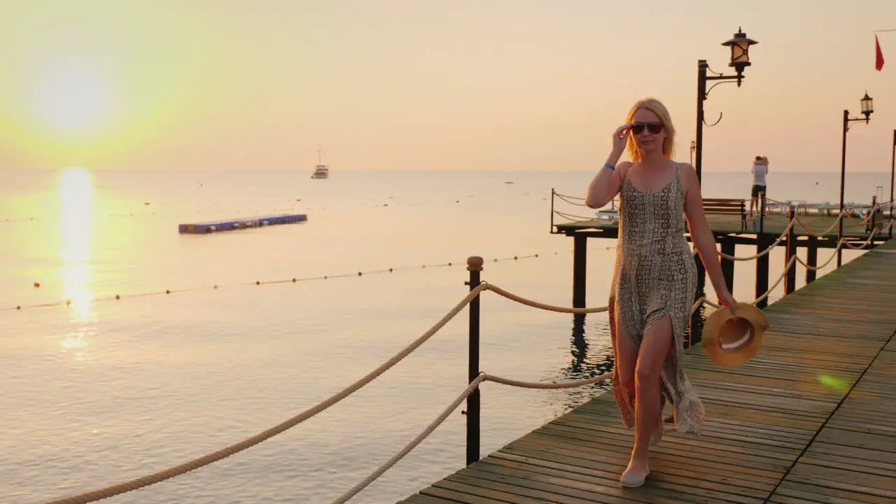 A Carefree Woman In A Light Dress With A Hat In Her Hand Walks Along The Pier