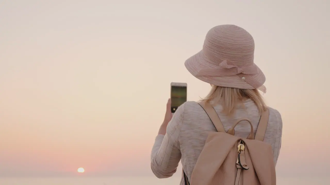 Young Stylish Girl In A Hat Takes Pictures Of The Sea And A Pink Sunrise On A Smartphone