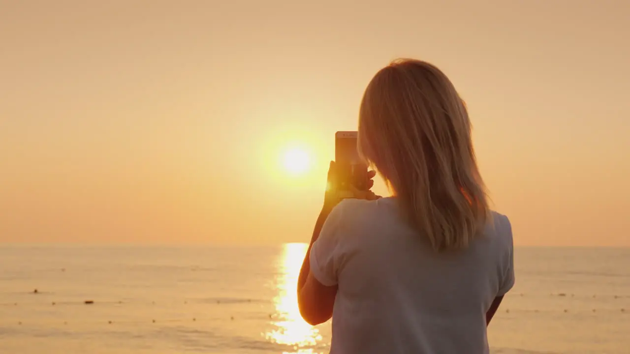 A Young Blonde Woman Is Photographing On A Smartphone A Pink Dawn By The Sea View From The Back