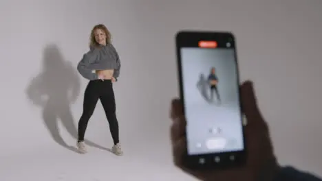 Person Recording Video On Mobile Phone Of Woman Doing Dance Practise On Grey Background In Studio