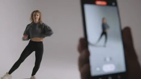 Person Recording Video On Mobile Phone Of Woman Doing Dance Practise On Grey Background In Studio 1