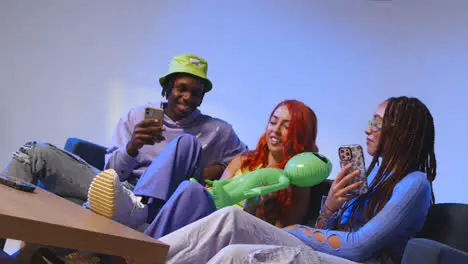 Studio Shot Of Group Of Young Gen Z Friends Sitting On Sofa With Toy Alien Gaming And Using Social Media On Mobile Phones