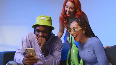 Studio Shot Of Young Gen Z Friends Sitting On Sofa Sharing Social Media Post On Mobile Phones 3