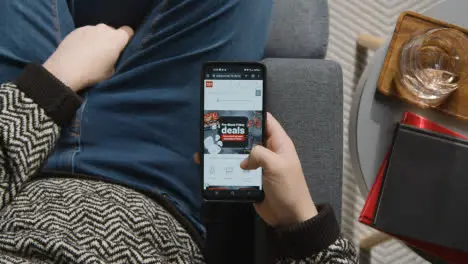 Overhead Shot Of Person At Home Shopping Online Looking At Argos Website On Mobile Phone On Black Friday