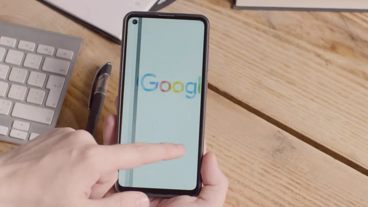 Person Holding Mobile Phone On Desk With Screen Showing Brand Logos For Apple Google McDonalds Microsoft Tesla Verizon And Visa