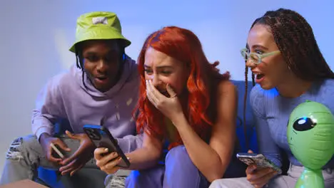 Studio Shot Of Young Gen Z Friends Sitting On Sofa Sharing Social Media Post On Mobile Phones