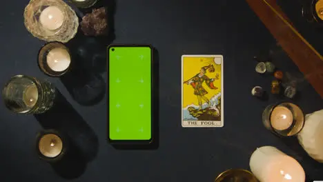 Overhead Shot Of Person Giving Tarot Card Reading With Green Screen Mobile Phone Next To The Fool Card On Table