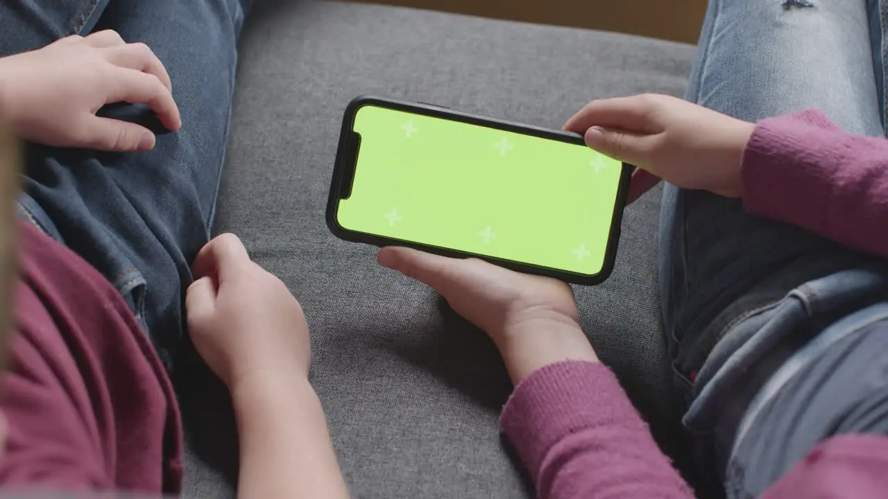 Close Up Of Two Children Sitting On Sofa At Home Looking At Green Screen Mobile Phone