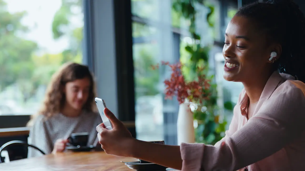 Young Businesswoman Wearing Wireless Earbuds Making Video Call On Mobile Phone In Coffee Shop