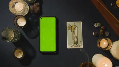 Overhead Shot Of Person Giving Tarot Card Reading With Green Screen Mobile Phone Next To The Hermit Card On Table