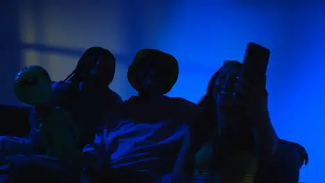 Studio Shot Of Group Of Gen Z Friends Sitting On Sofa Posing For Selfie On Mobile Phone At Night With Flashing Light