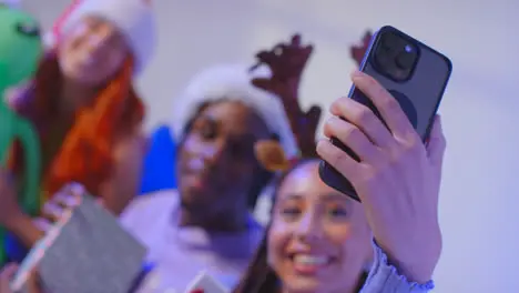 Studio Shot Of Gen Z Friends At Christmas Sitting On Sofa Wearing Santa Hat And Reindeer Antlers Taking Selfie On Mobile Phone 2