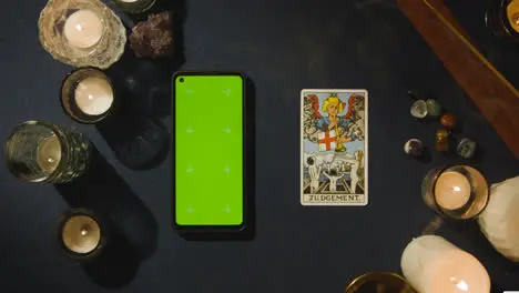 Overhead Shot Of Person Giving Tarot Card Reading With Green Screen Mobile Phone Next To Judgement Card On Table