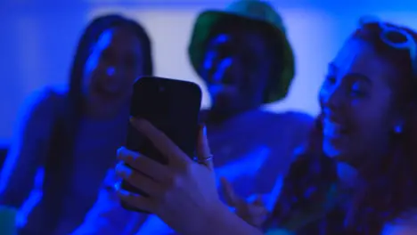 Studio Shot Of Group Of Gen Z Friends Sitting On Sofa Posing For Selfie On Mobile Phone At Night With Flashing Light 4