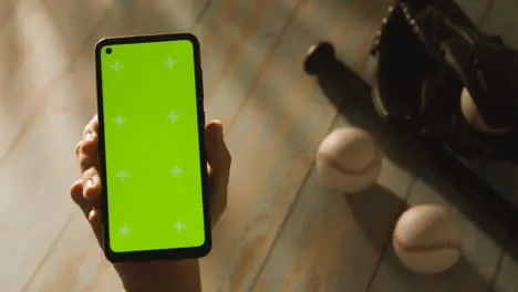 Person Holding Green Screen Mobile Phone Above Baseball Still Life With Bat Ball And Catchers Mitt On Aged Wooden Floor