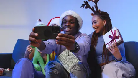 Studio Shot Of Gen Z Friends At Christmas Sitting On Sofa Wearing Santa Hat And Reindeer Antlers Taking Selfie On Mobile Phone 1