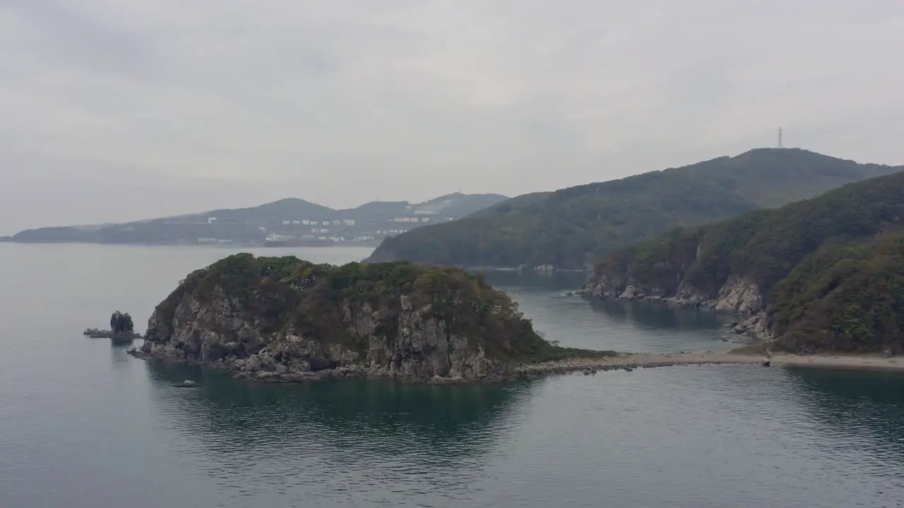 Sea coastline with island cliffs with green trees on the sides on an overcast day with port facilities in far distance