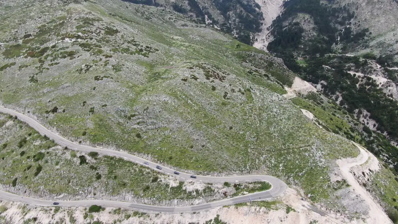 Drone view in Albania flying over a green and rocky mountain with serpent road climbing a port with cars driving down on a cloudy day