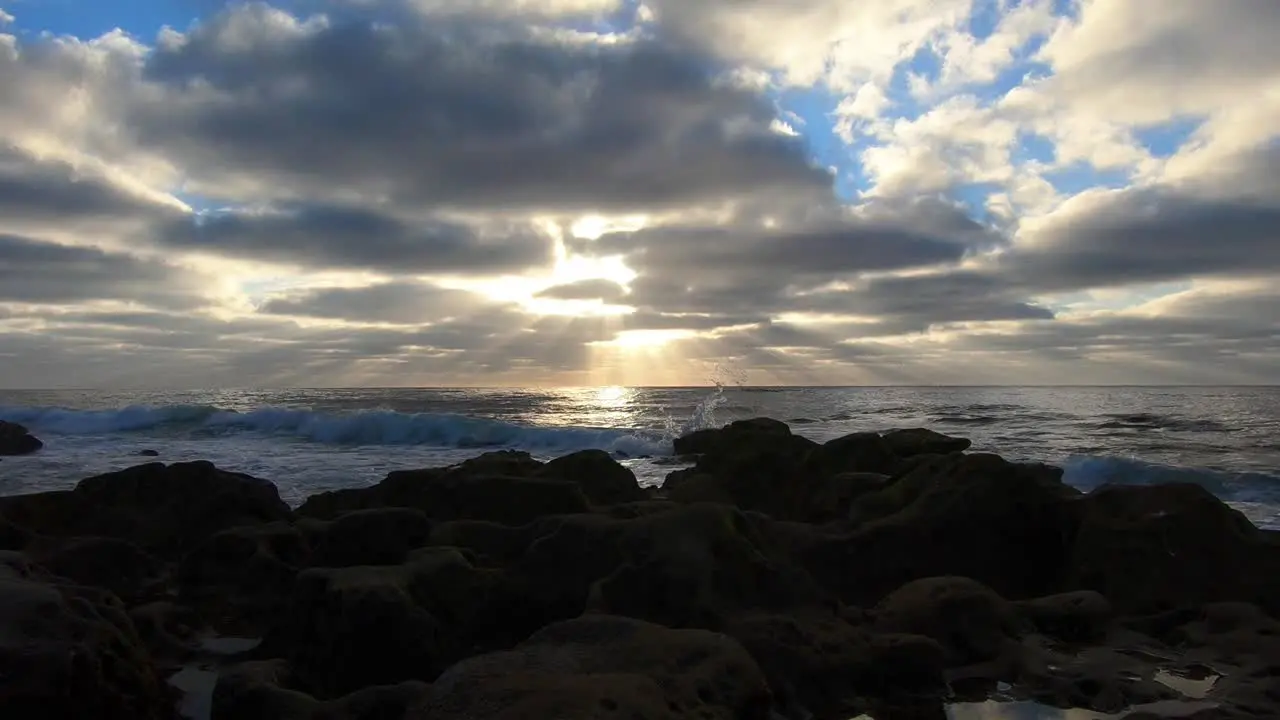 establishing shot of sun rays peaking through the clouds over the ocean on a cloudy day with small waves rolling onto shore with rocks in the foreground and patches of blue sky