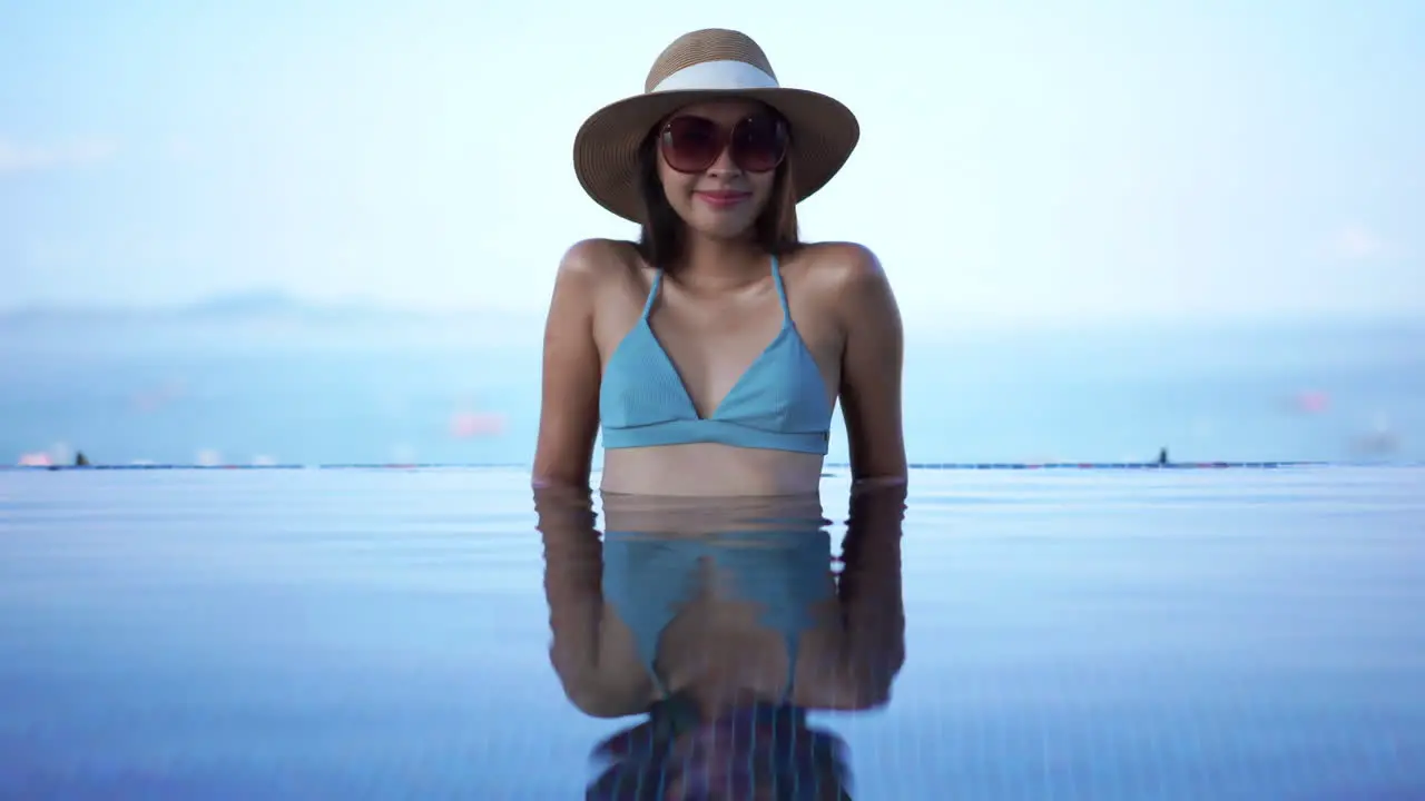 Beautiful Asian woman in bikini wearing sun hat and sunglasses sitting in swimming pool