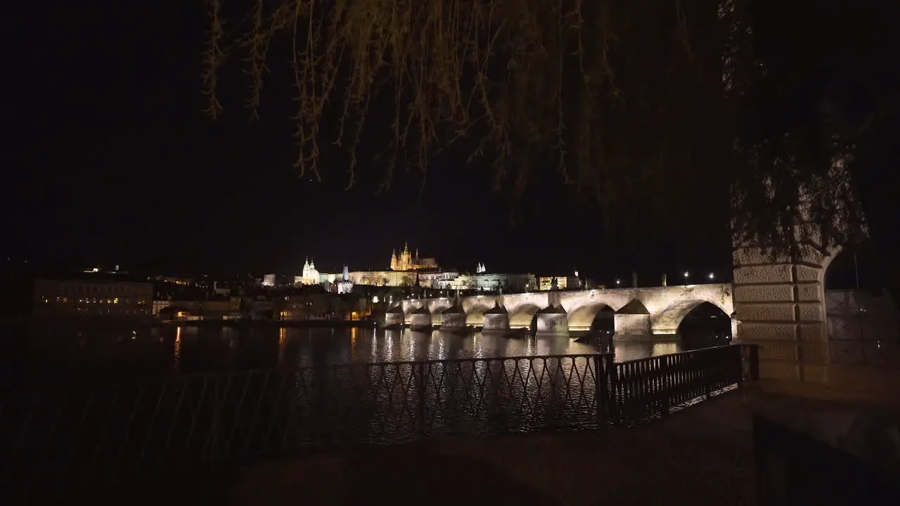 The Prague Castle and Charles bridge over river Vltava in the historical centre of Prague Czechia lit by lights at night viewed from the other side of the river pedestal shot from under a willow tree