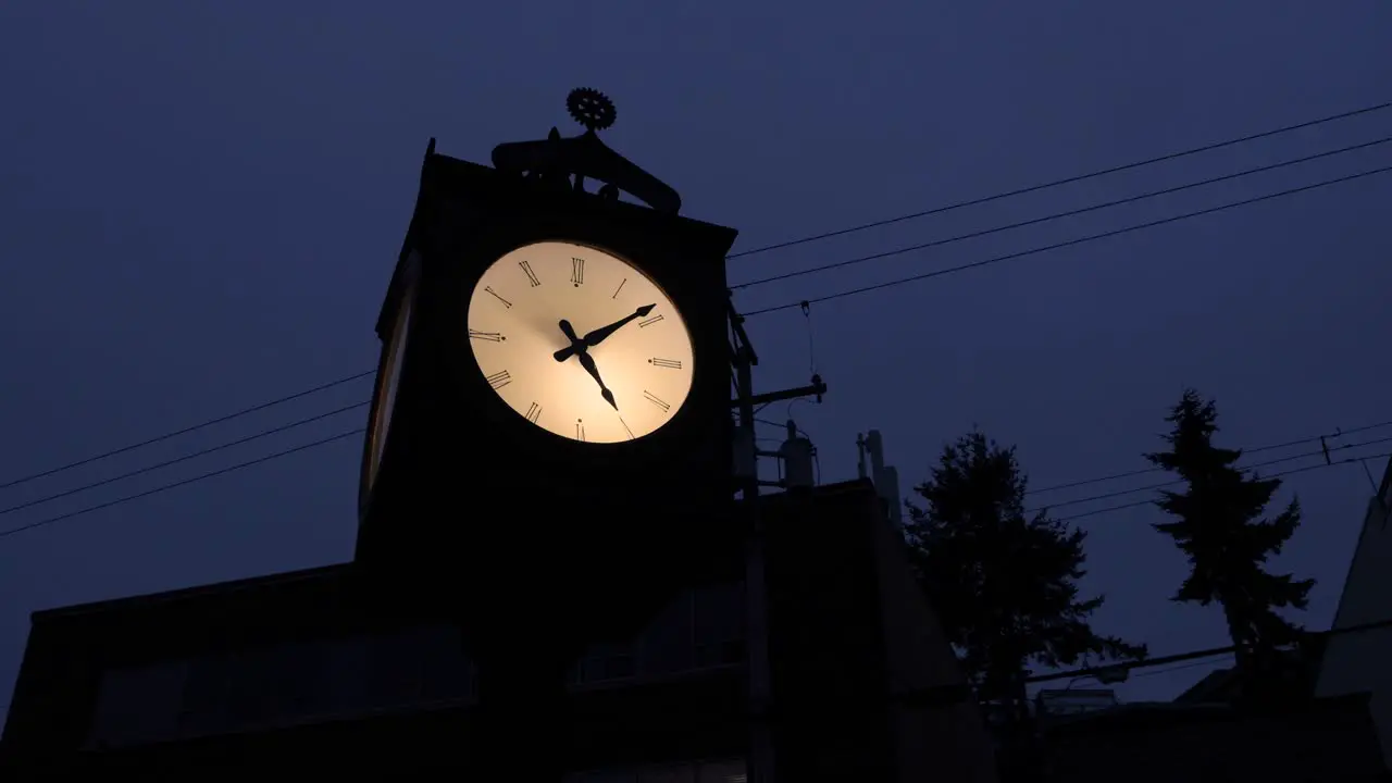 Silhouette of an old Illuminated grandfather clock on a dark evening amongst buildings and electrical wires trees sky