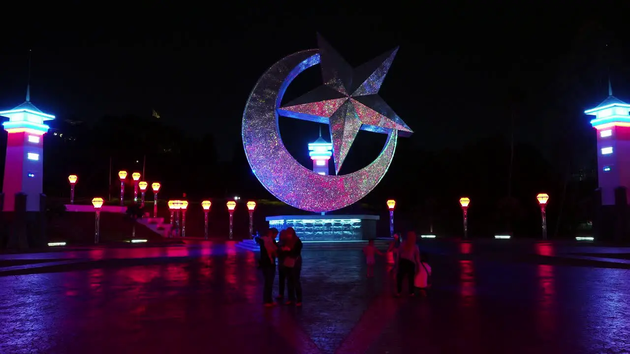 Star and crescent statue lit up by colorful lights as tourists take pictures beneath it on a plaza at night