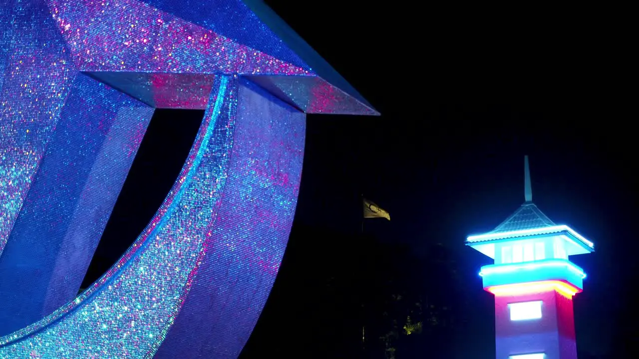 Corner of glittery star and crescent statue lit by colorful lights at night with a flag flying in the night in the distance