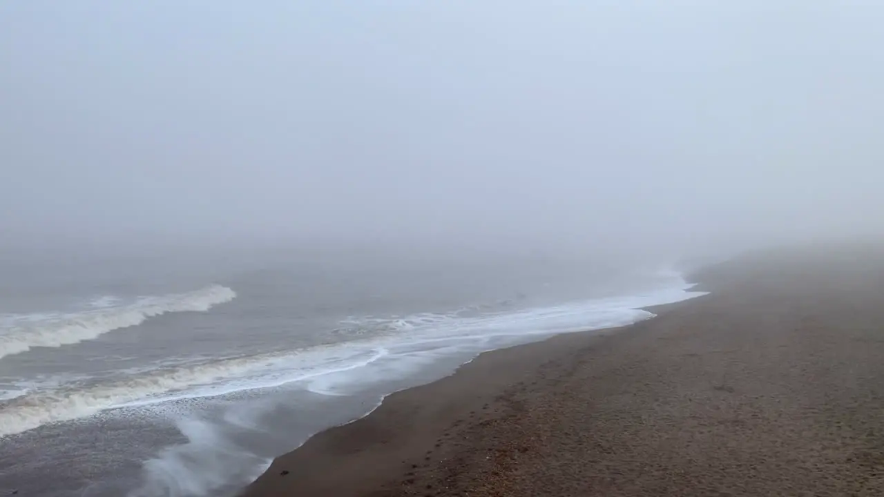 A misty coastline with waves rolling in to the beach shore beneath a foggy grey sky