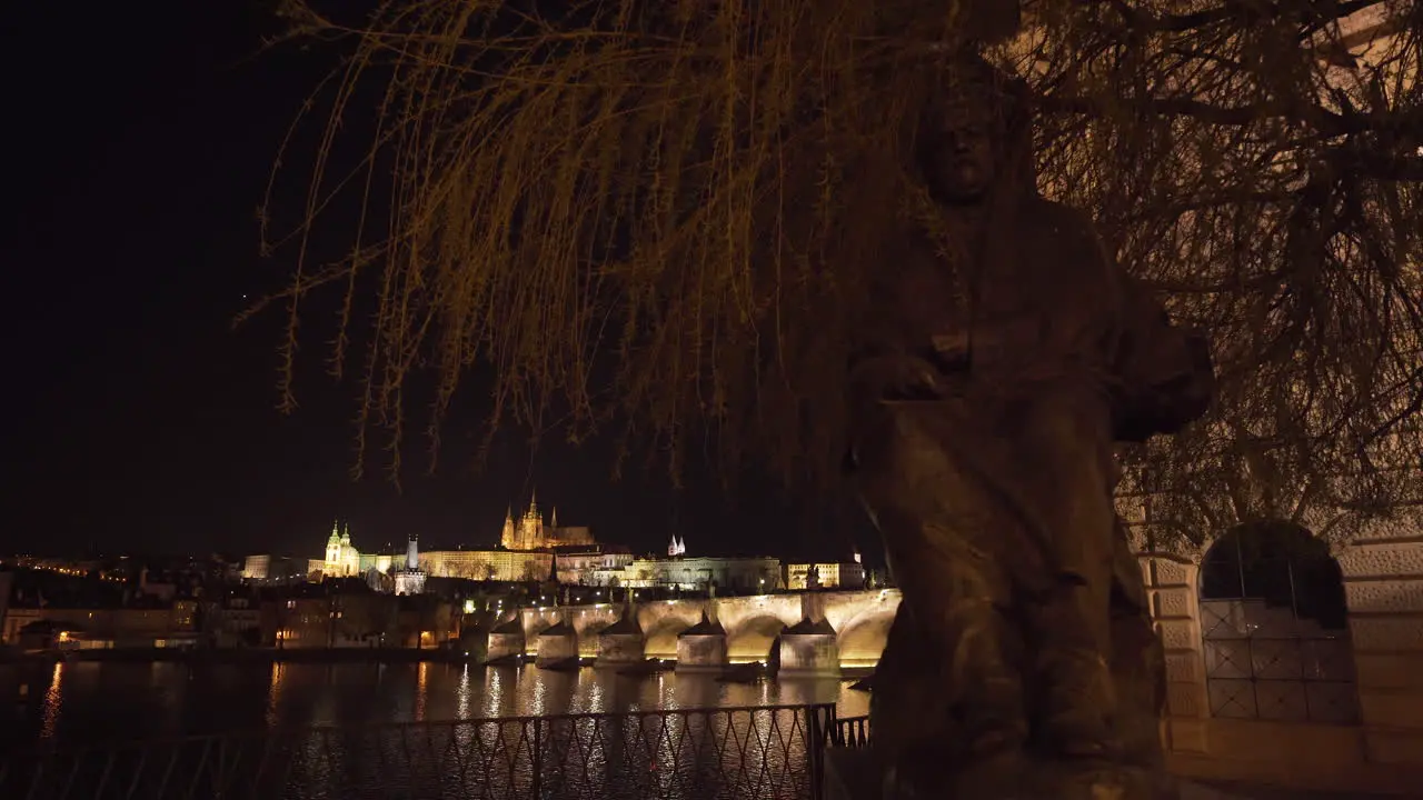 The Prague Castle and Charles bridge over river Vltava in the historical centre of Prague Czechia lit by lights at night shot from under a statue of a king and a tree on the other side of the river