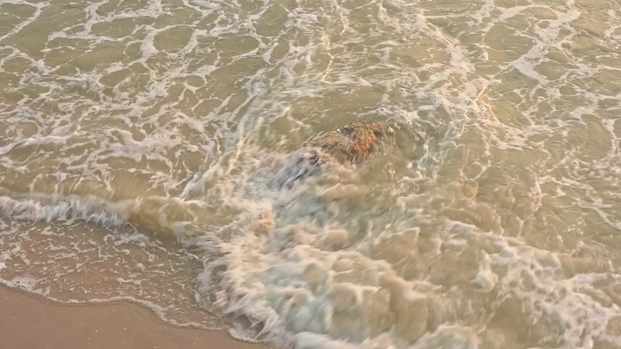 Slow-motion of surf rushes in to cover a large moss-covered rock in the middle of a beach