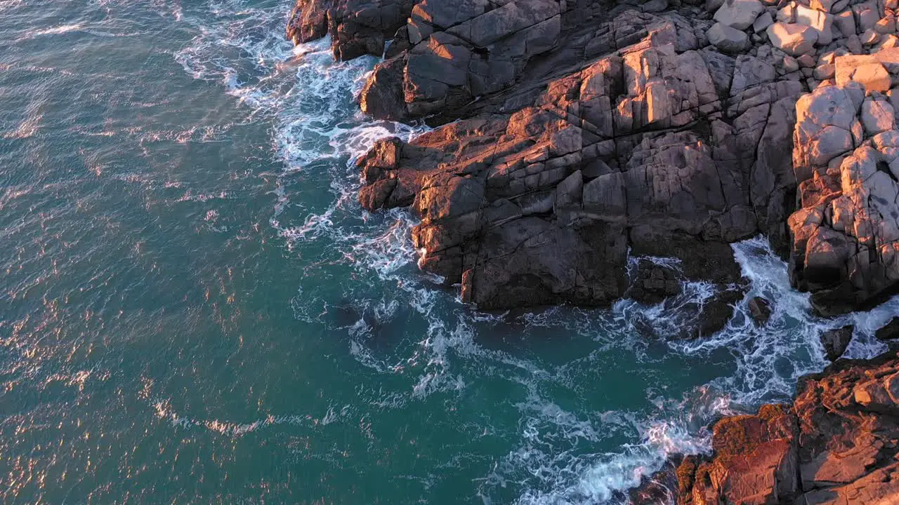 Aerial TOP DOWN of the waves crashing on the rocks below Nubble Lighthouse in Maine lit by a cold December sunrise